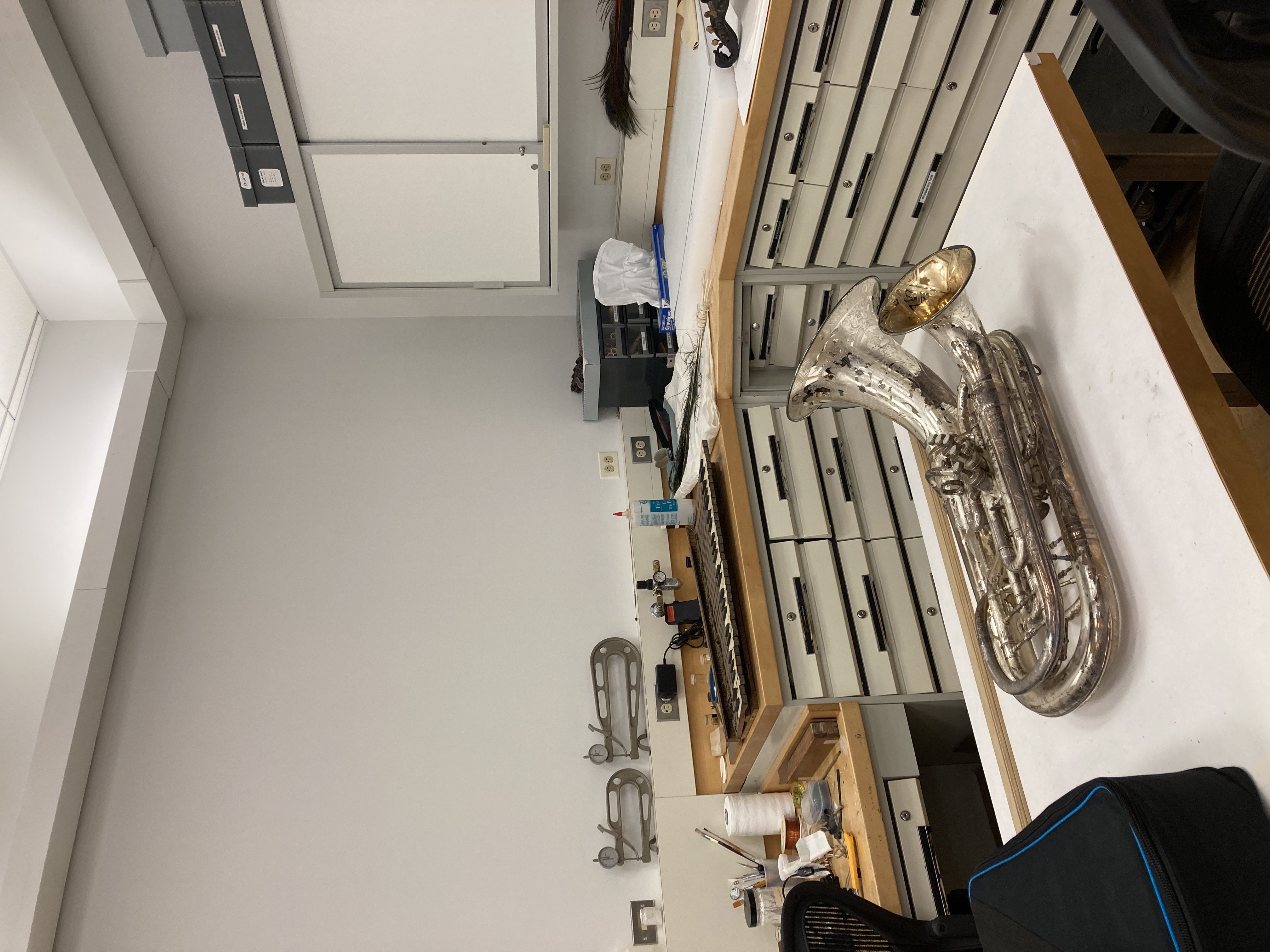 A photo of an antique tuba in the museum. It is on a table with more antiques in the background.