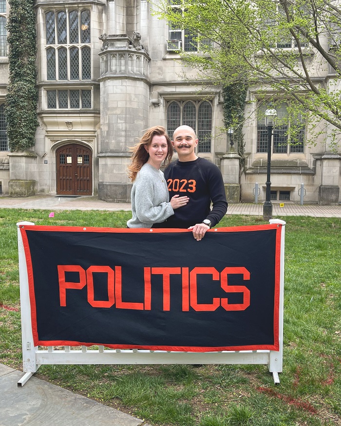 Xander with his partner Jenna standing on campus behind a black and orange sign that says "Politics"